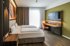 Hotelzimmer mit zwei Einzelbetten, Schreibtisch, großem TV und heller Fensterfront.