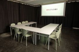 Konferenzbereich mit U-förmig gestellten weißen Tischen, hellen Stühlen und großer Projektionsleinwand mit VFX-Studios-Logo.