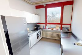 Helle Gemeinschaftsküche mit großem Edelstahlkühlschrank, weißen Einbaumöbeln, Fensterfront und kompletter Kochzeile.